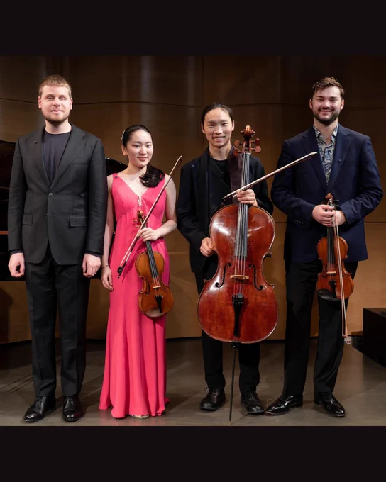 Four musicians are standing and smiling in front of a wooden backdrop. They are dressed in formal attire, with one holding a violin, one holding a viola, and one holding a cello. A grand piano is partially visible behind them.