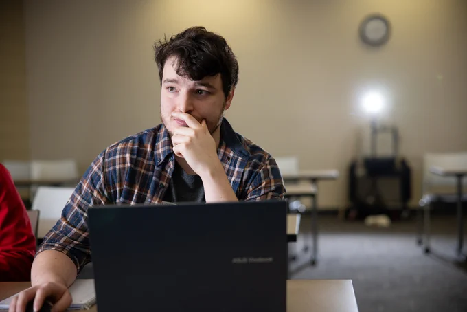 A person sitting at a table with a laptop, appearing thoughtful. 