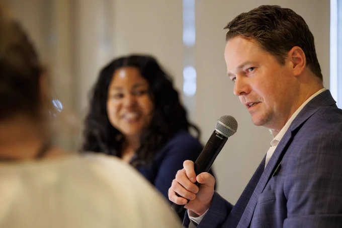 Photo of man holding a microphone and speaking. There is a woman in the background listening. 