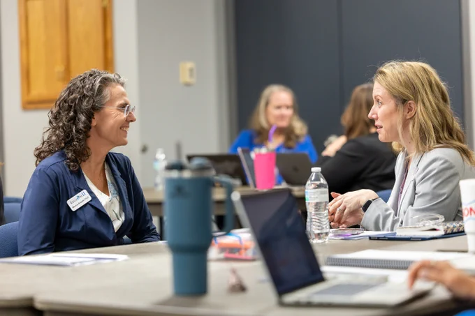 Two individuals are seated and engaged in conversation at a conference table. The table has laptops, water bottles, and notebooks. Other people are visible in the background, also interacting.