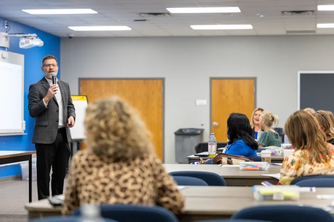 Nick Keith is speaking into a microphone at the front of a conference room. Several people are seated at tables, attentively listening. The room has gray walls and a whiteboard at the front.