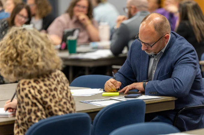 A person in a blue blazer is writing on a notepad at a conference table, surrounded by others engaged in discussion.