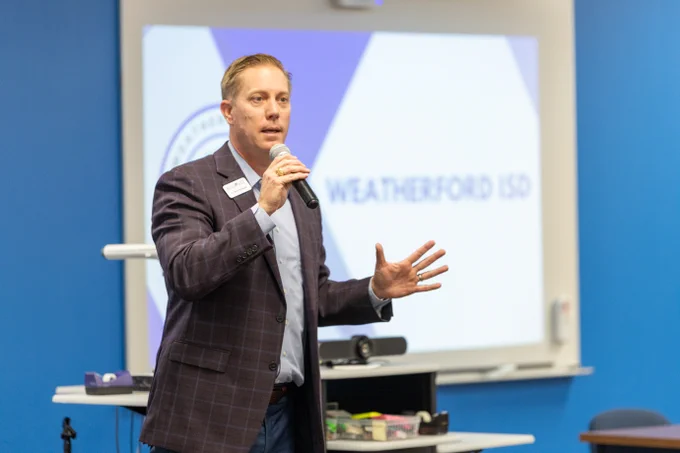 Dr. Brad Schnautz is speaking into a microphone at the CAN meeting. The background features a large screen displaying a presentation with the text "Weatherford ISD." Dr. Schnautz is wearing a checkered suit and holding the microphone with one hand while gesturing with the other.
