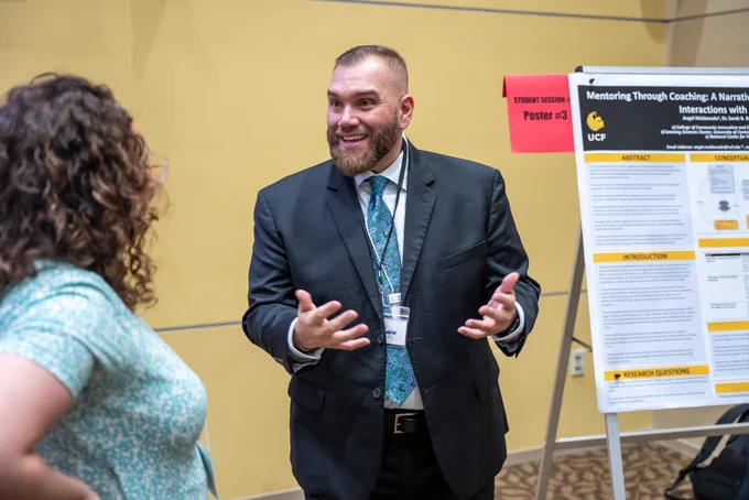 Angel Maldonado standing next to his research poster presentation and smiling, hands slightly raised in animated conversation with a person to his right.
