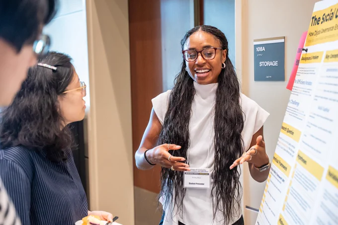A student smiling, hands slightly raised in animated conversation, while presenting her research poster to two people listening on her right.