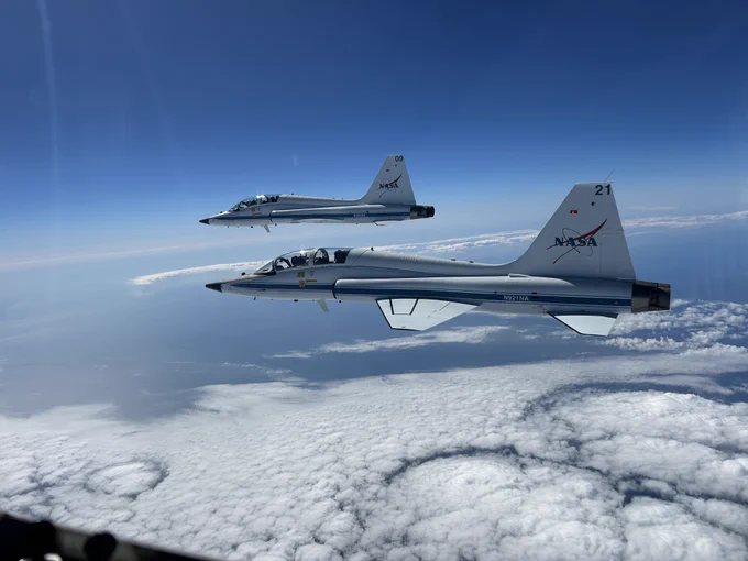 Two T-38 planes fly in the air. Clouds can be seen underneath the planes. The sky is blue in the background. Credit: NASA