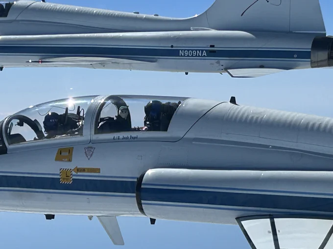 One of the Artemis II astronauts holds up the \u201CRise\u201D zero gravity indicator doll from the window of a T-38 plane flying in mid-air. The sky is blue in the background. Credit: NASA