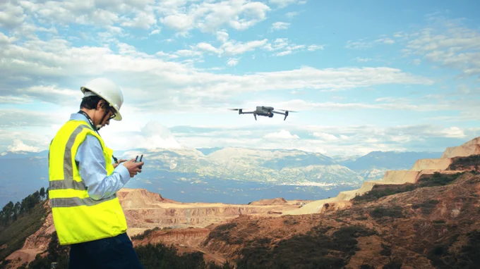 A person wearing a hard hat and a neon safety vest operates a drone using a remote control at a hilly mining site under a partly cloudy sky.
