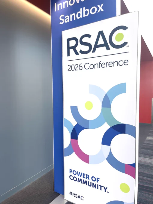 Signage for RSAC 2026 Conference featuring colorful abstract circular designs and the phrase "Power of Community" with the hashtag #RSAC. The banner is positioned next to a tall display labeled "Innovation Sandbox." The surroundings include a blue and red wall and a gray carpeted floor.