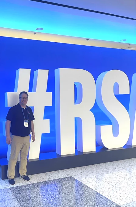 A person stands beside large white letters that read "#RSA" in a brightly lit area. The backdrop has a blue gradient, and the person is wearing a conference badge.
