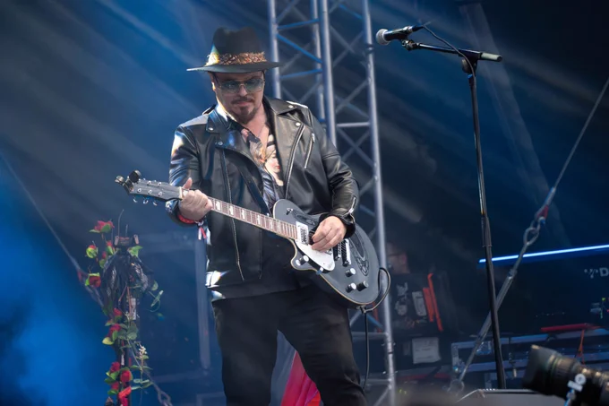 A man wearing black jeans, a black leather jacket and black hat. He is playing a black electric guitar and there is a microphone in front of him.
