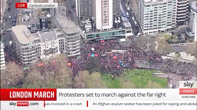An aerial news broadcast shot from Sky News showing a large crowd of protesters gathered in a public square in Central London. The crowd is densely packed between high-rise city buildings and a green park area. A red and white news banner at the bottom reads, "LONDON MARCH: Protesters set to march against the far right." Small colorful tents and banners are visible within the crowd.