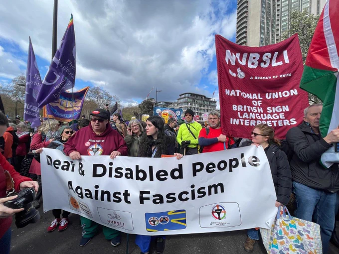 Image shows deaf and disabled people gathered behind a white banner that says deaf and disabled people against fascism in the background Is a red banner that national union british sign language interpreters
