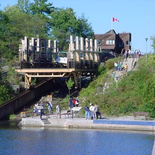 Lock and marine railway structure beside a body of water, with a large wooden framework and rails descending into the canal. Several people stand on a viewing platform near the water and along a path that climbs up a hill to the right. At the top of the hill, a building with a Canadian flag is visible among trees. The area is surrounded by green vegetation under a clear sky.
