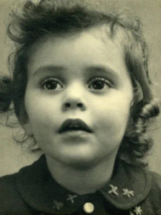 Photograph of the face of a few-year-old girl. She has wavy hair with curls on the sides. Under her neck she has a button-down shirt with a semi-circular collar with a pattern of flowers on it. She is looking slightly upwards and has a slightly open mouth.