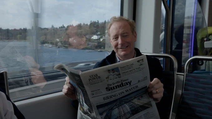 Brad Smith sits on a light rail train reading The Seattle Times Sunday edition. Through the window beside them, water and a tree-lined shoreline are visible, as the train travels along I-90 over Lake Washington.