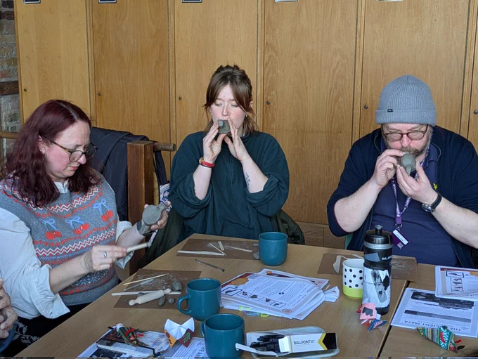 Three people working with clay. Two are trying to make sounds by blowing through the clay