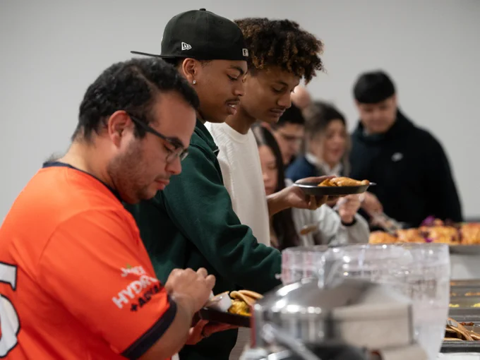 A group of people serving themselves food from a buffet table. One person is wearing an orange jersey with the a logo.