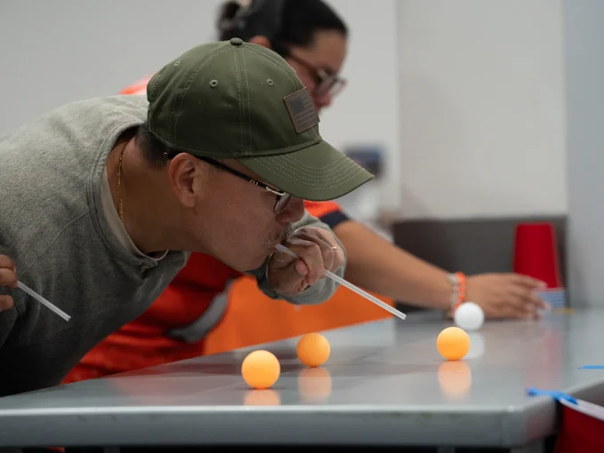 Two people are engaged in a game using straws to blow table tennis balls across a table. Three orange balls and one white ball are visible. There are red plastic cups in the background.