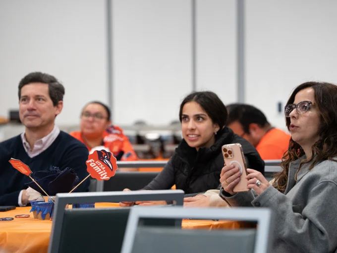Group of people seated at tables with orange tablecloths, focused on a presentation. Some are holding phones. A few decorative items and papers are on the tables.