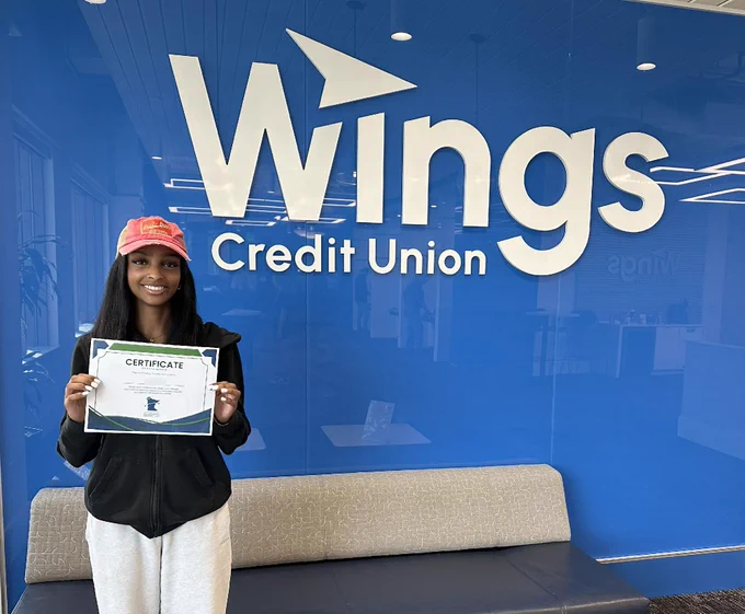 A person is standing in front of a blue wall with the Wings Credit Union logo. They are holding a certificate and wearing a red cap. There's a couch in the foreground.