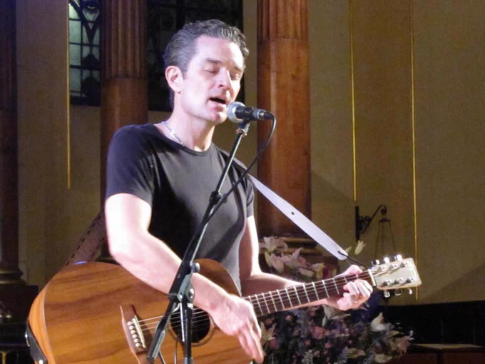 The image shows a musician performing indoors. He is playing an acoustic guitar and singing into a microphone with his eyes closed. He is wearing a black T-shirt and has short, dark hair. The guitar is wooden with a visible grain pattern. Behind him, there are tall, wooden columns with ornate detailing near the top. The background also includes stained glass windows and floral arrangements. The lighting casts a warm glow, highlighting the performer and adding depth to the background.