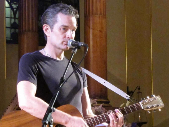 The image shows a man on stage, playing an acoustic guitar and singing into a microphone looking out into the audience. He wears a black t-shirt and a light-colored guitar strap across his shoulder. The background reveals tall wooden pillars and stained glass windows, indicating a setting that resembles a church or hall. The lighting focuses on the performer, creating a contrast with the darker surroundings.