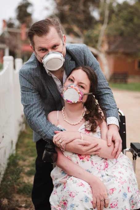 A white woman in her wheelchair wearing her Flo mask with her partner wrapped around behind her wearing his Flo mask.