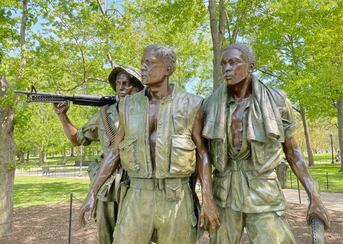 Three bronze statues of Vietnam War era American soldiers wearing jungle fatigues, standing together among trees at the Vietnam Veterans Memorial. 