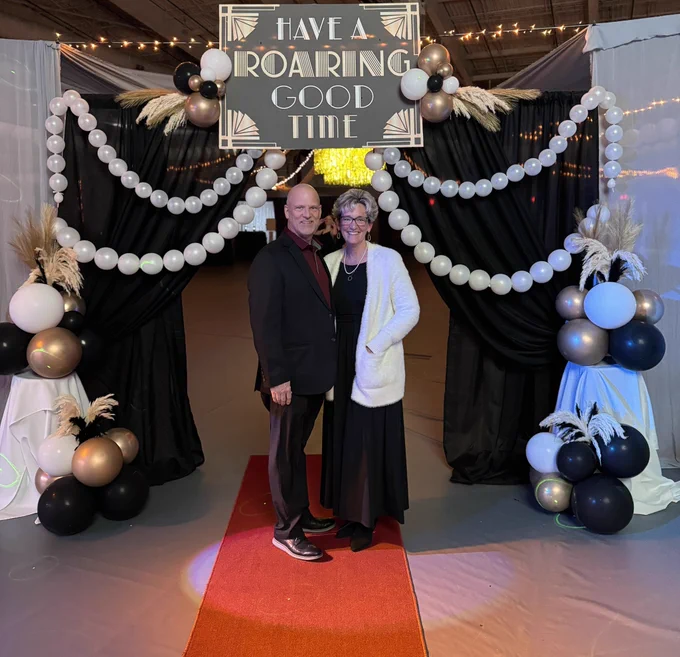 Superintendent and husband smiling at a high school prom as they stand under a sign that says, “Have a roaring good time.”