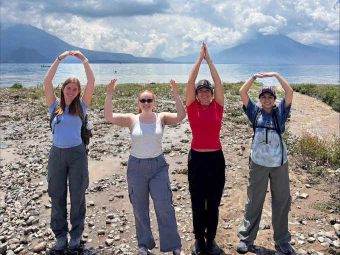 Four people stand on a rocky shore, forming the letters "OHIO" with their arms. They are near a lake with mountains in the background under a partly cloudy sky. They appear to be enjoying an outdoor activity.