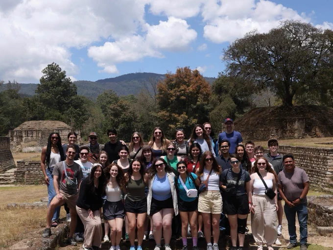 A group of people posing together outdoors in front of ancient ruins with mountainous scenery in the background. The sky is partly cloudy and trees surround the area.