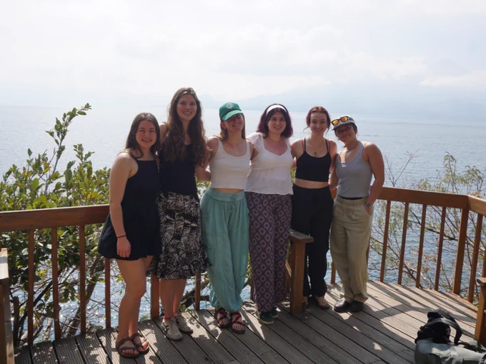 A group of six individuals stands on a wooden deck overlooking a large body of water, with foliage in the background. They are casually dressed and smiling, enjoying a sunny day outdoors.