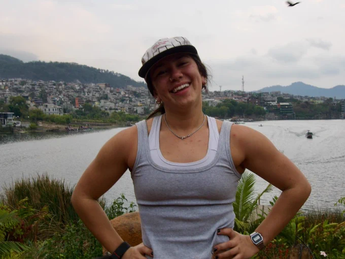 A person stands smiling near a body of water with a town and hills in the background. They are wearing a cap, sleeveless top, and watch. A bird is flying in the sky. The atmosphere is overcast.