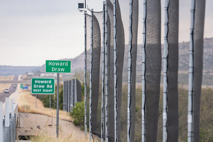 A roadside view shows signs for "Howard Draw" and "Howard Draw Rd Next Right" on a highway. Several tall, black mesh barriers run parallel to the road, with a hilly landscape in the background.