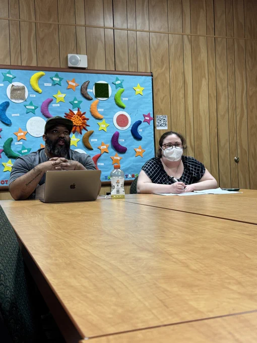 A Black man in a hat and a white woman wearing glasses and a mask sit at the front of a table speaking