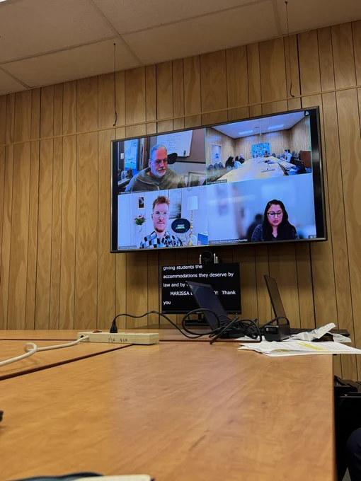 A big television screen shows 2 men and 1 woman on a Zoom screen talking.