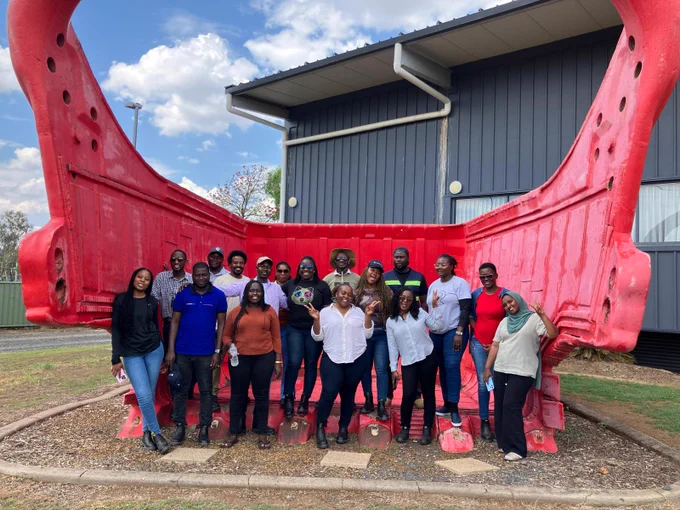 Group photo of participants posing cheerfully inside a large red industrial excavator bucket outdoors on a sunny day.