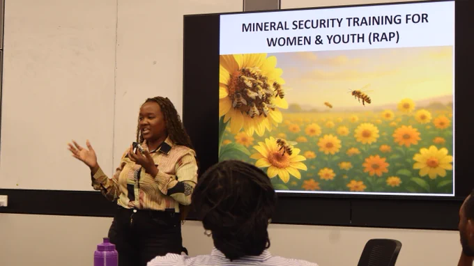 Participant presenting at the front of the room into a handheld microphone, with a slide to their right showing flowers and bees with the title "mineral security training for woman and your". 
