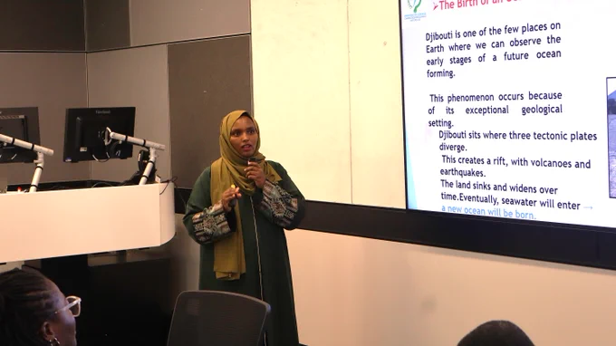 Participant presenting in green attire explains geological phenomena with a slide about Djibouti's tectonic activity and ocean formation.