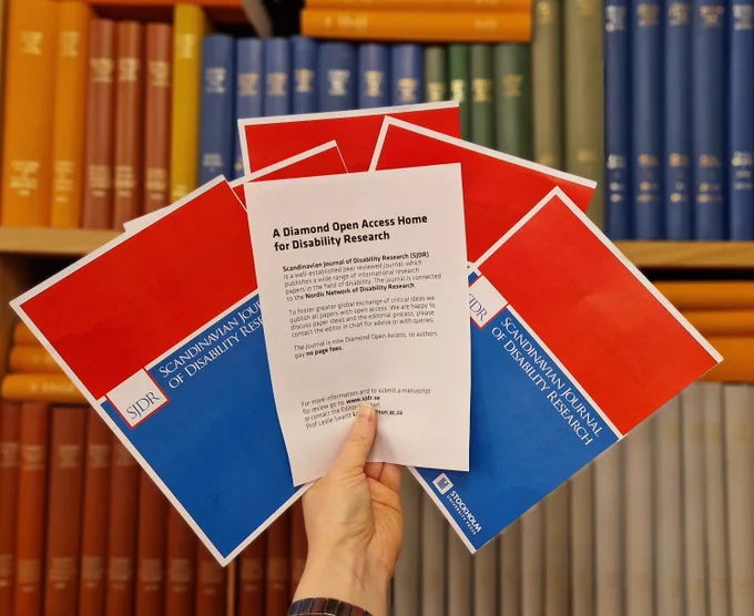 A hand holds two-sided flyers in front of a bookshelf. One side of the flyers has the information about Diamond Open Access, the other has the graphics of the journal.