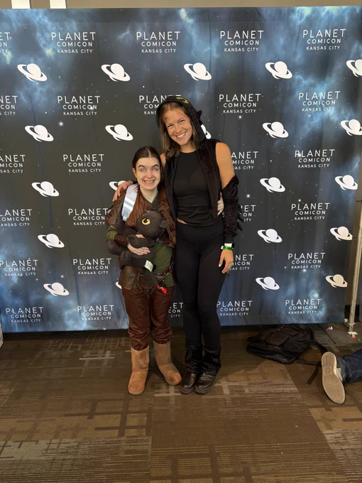 Hiccup and Toothless standing in front of the Planet Comicon Kansas City backdrop! I'm in my Hiccup cosplay, and my friend borrowed my Toothless pieces so she could be Toothless! It worked out because I had to hold onto her elbow known as sighted guide, so in a way I had a guide dragon!