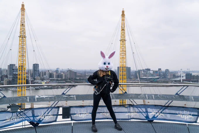A person stands on top of the O2 dressed in black with a white bunny head on. London's skyline is in the background
