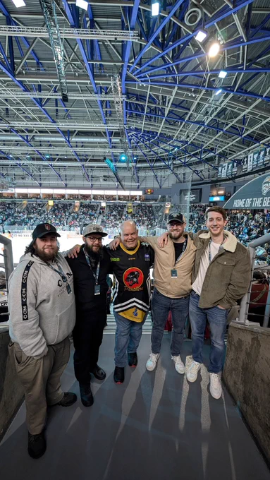 5 commentators stood in a semi-circle wit their backs to the Ice Arena, Belfast. Their arm around shoulders, smiling.