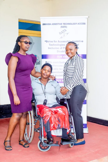 Two women standing and smiling while posing with a third woman who is seated in a wheelchair in front of a RATA banner.