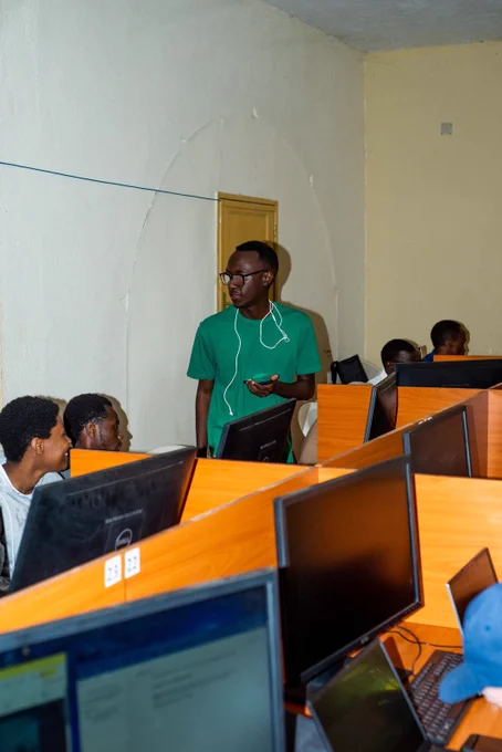A man in a green t-shirt stands in a computer lab, looking toward students seated at wooden desks with monitors.