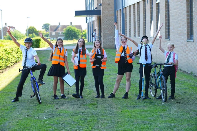 Group of six schoolchildren in uniforms and orange safety vests cheer outdoors near a school building, two with bicycles. 