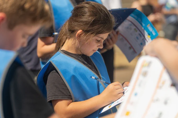 Child wearing a blue vest intently writing on a clipboard during an outdoor educational activity.