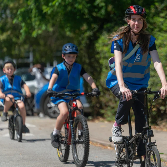 Group of children wearing helmets and blue safety vests riding bicycles on a sunny road surrounded by greenery.