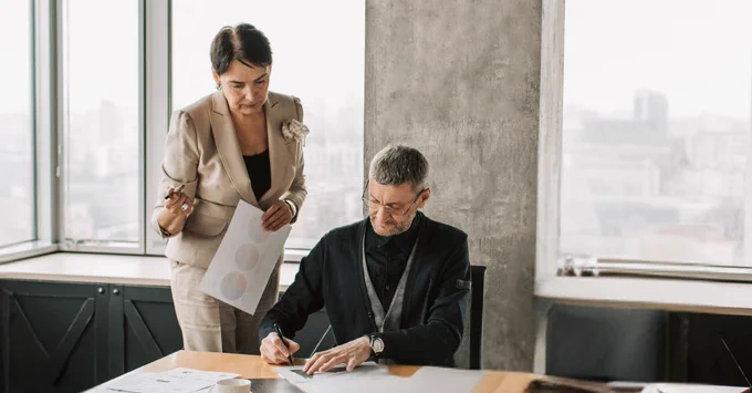 Two colleagues collaborate on paperwork in a modern office. One is standing by the desk while the other is seated and writing.
